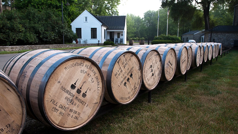 Bourbon barrels on the grass with a small white house in the background among trees 